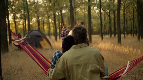 Rear-view-of-a-happy-couple,-a-brunette-man-and-a-girl-in-a-black-hat,-sitting-on-a-red-hammock-near-the-rest-of-the-company-during-a-camping-stop-in-a-sunny-summer-forest