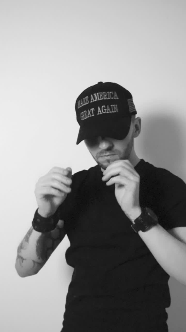 Grayscale footage of a man with tattoos, short hair and a beard putting on a dark black MAGA Make America Great again hat, crossing his arms and begins to nod with approval, showing he likes Trump.