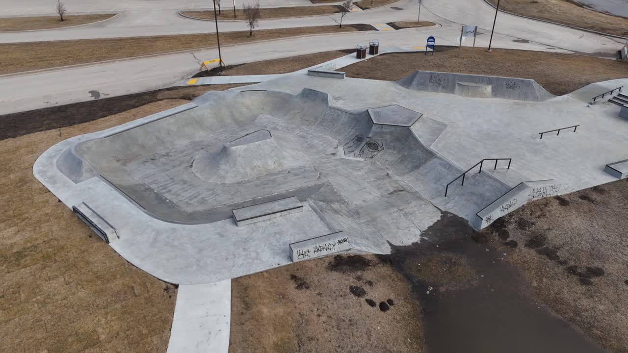 Drone che orbita attorno a uno skatepark in cemento di nuova costruzione, mostrando ringhiere, rampe, erba primaverile marrone e acqua in un fosso vicino sotto cieli sereni.