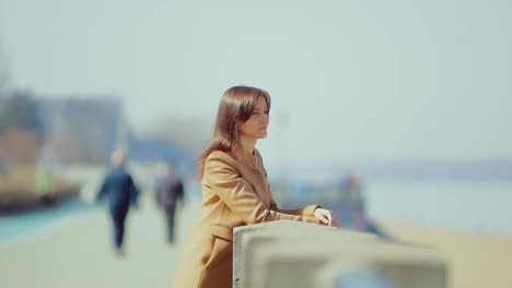 young business lady is standing at the parapet on the embankment and dreaming. people walk by the river in early spring. circular motion of the camera.