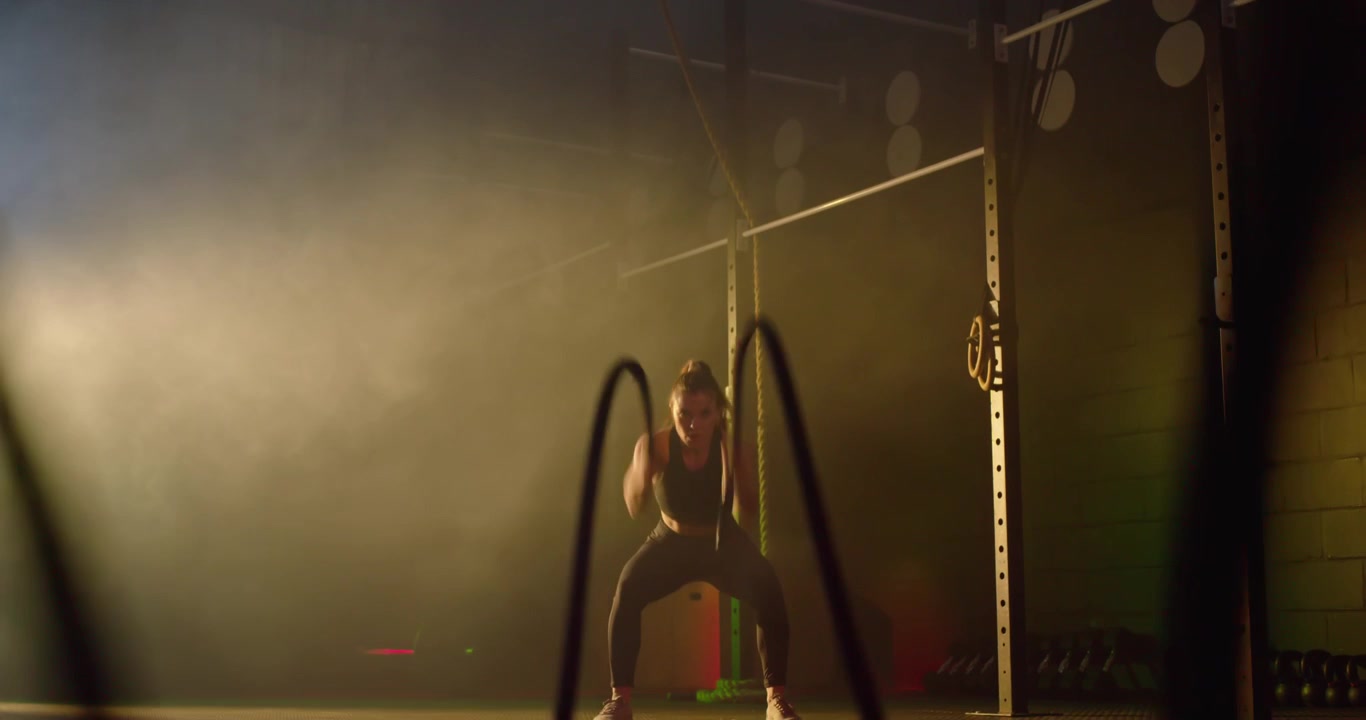 Donna afroamericana che entra nell'inquadratura, afferrando le battle ropes, facendo squat e allenandosi in reggiseno sportivo.