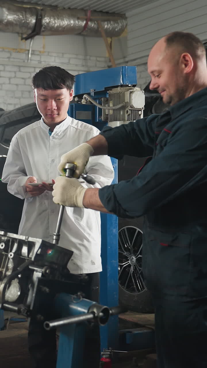 Mechanic instructing students on engine repair in an automotive workshop, student in lab coat observes while holding tablet, another student listens attentively, background includes tools and car