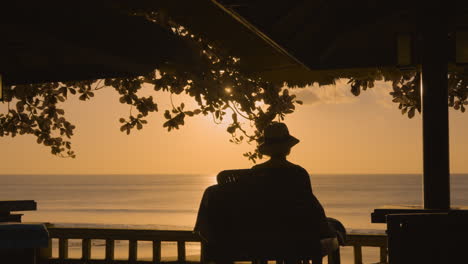 Silhouette-of-a-woman-in-a-wide-brim-hat-relaxing-while-watching-a-breathtaking-sunset-at-a-beach-resort,-medium-orbit-and-pan-left