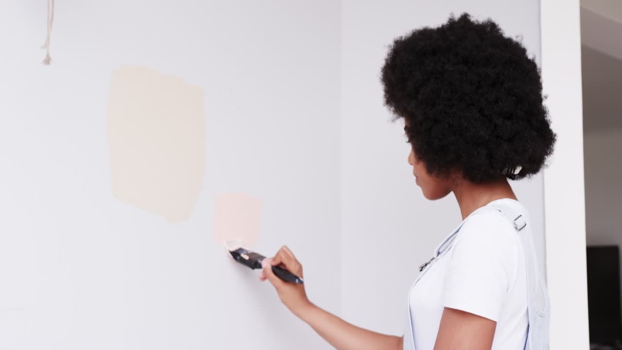 Donna afroamericana in tuta da lavoro che applica un campione color pesca con un pennello in una stanza, valutando, con spazio per il testo.