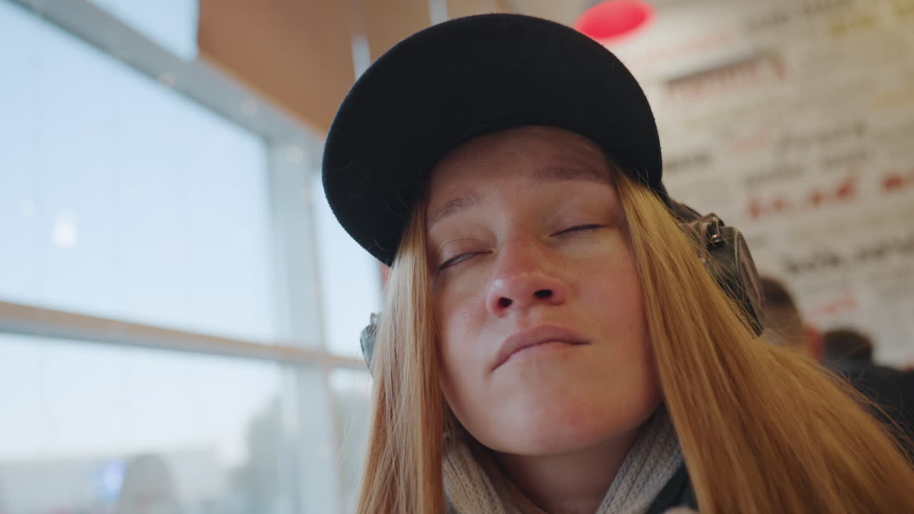 Maskeret kvinde ved vindue kigger ud over byens skyline, hovedtelefoner og kasket, strikket tørklæde og blød solbelyst glød, transitlounge-baggrund med fjerne passagerer, indadvendt udtryk, der skifter mellem ro.