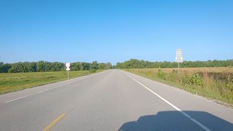 POV-driving-past-trees-and-fields-on-a-rural-paved-road-in-late-summer-in-Iowa,-USA