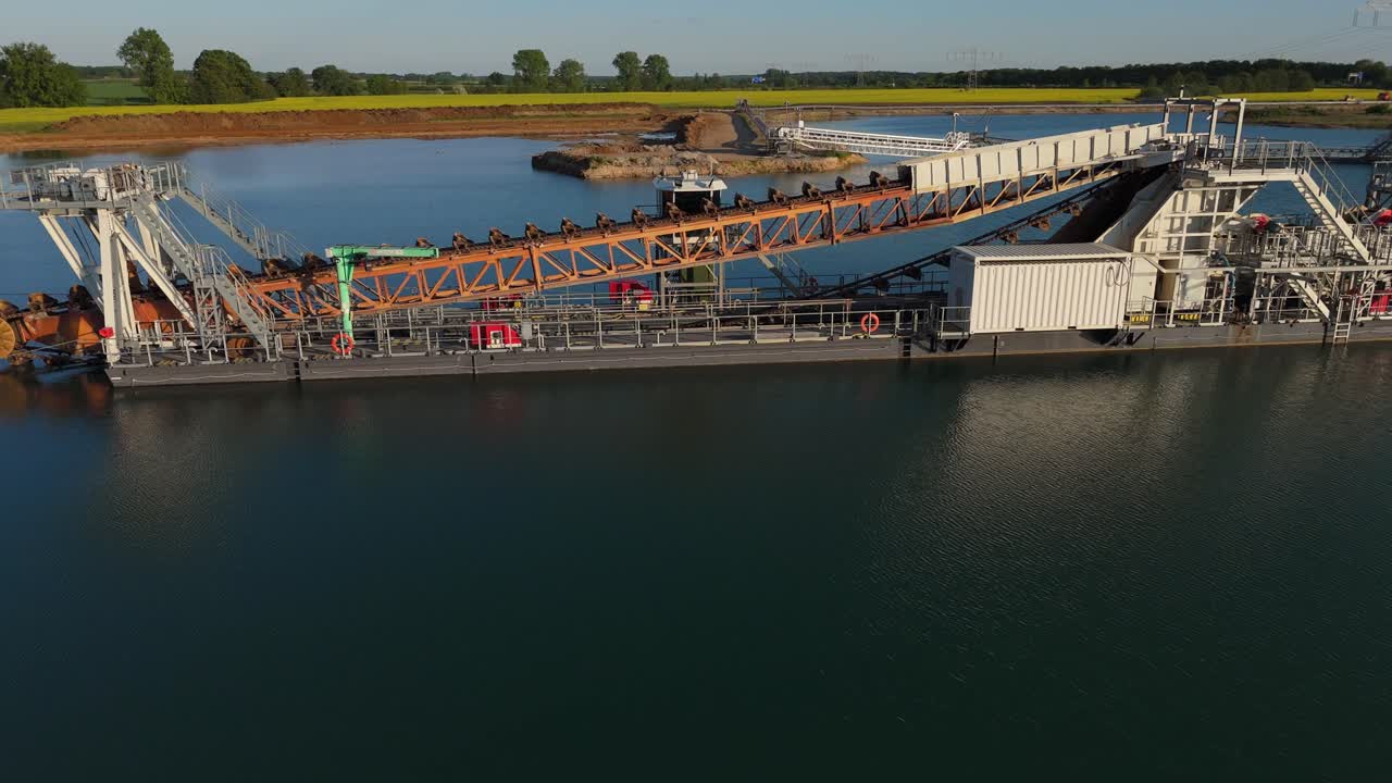 Drone footage of a floating dredger with conveyor belt operating on a gravel lake near Leipzig, Germany. The industrial scene is framed by calm water and early summer fields in the background.