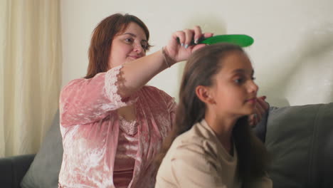 Mother brushing daughter’s hair on couch in cozy living room at home. Intimate, peaceful family moment with soft natural light coming through the window, creating calm and loving atmosphere