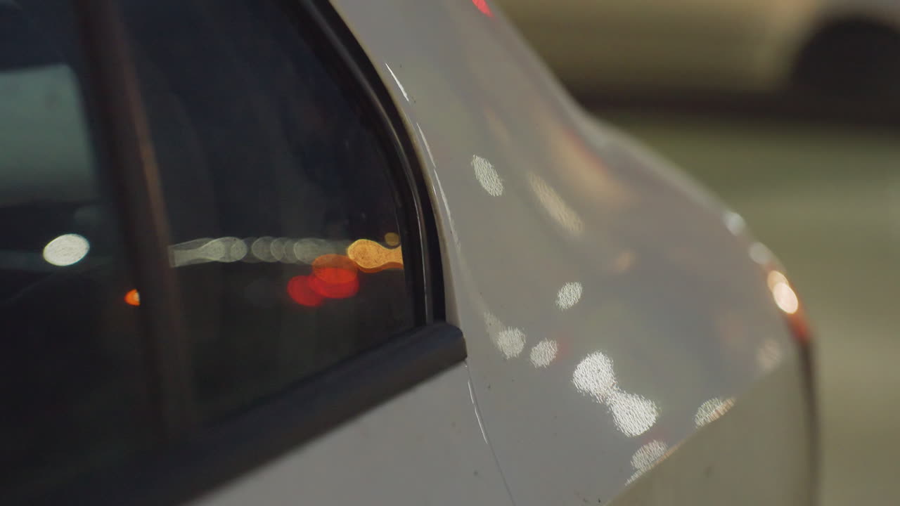 Close up side view of ash color car parked under streetlights at night, with vehicle window reflecting passing cars and blurred city lights creating glowing bokeh effect on body surface