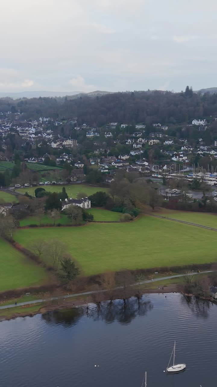 A vertical drone pan shot over Lake Windermere towards Bowness, passing above small marina. Winter trees and Bowness town are in the foreground as hilltops glow under sunlight breaking through clouds