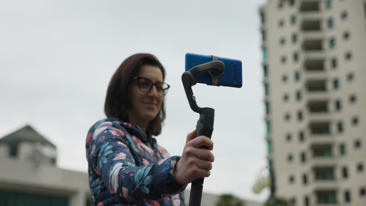 mujer adulta con gafas sostiene estabilizador de cardan con teléfono móvil azul en modo retrato