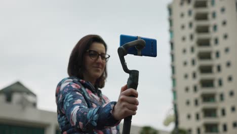 adult woman with glasses holds gimbal stabilizer with blue mobile phone in portrait mode