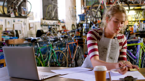 -Woman-writing-on-note-pad-while-using-laptop-at-workshop-4k