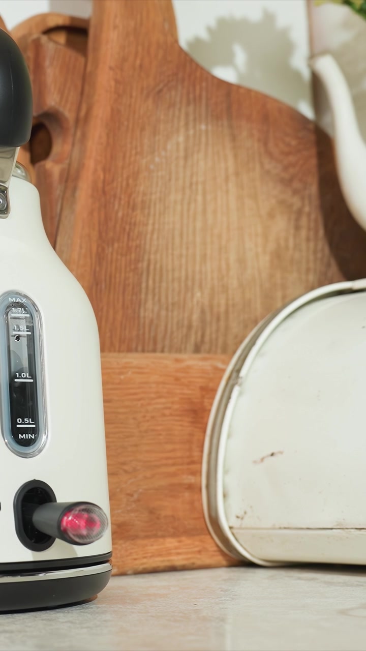 Close up of white electric kettle boiling water. Kitchen countertop features wooden cutting boards metal tin. Camera captures slight shift to reveal more kitchen items