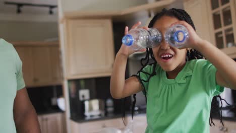 Glücklicher-Afroamerikanischer-Vater-Und-Tochter,-Die-In-Der-Küche-Mit-Plastikflaschen-Zum-Recycling-Spielen