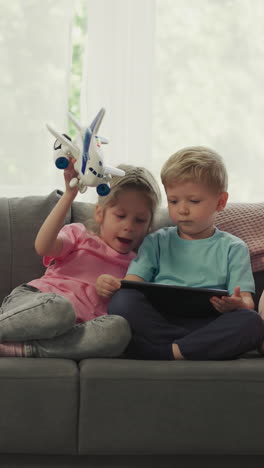 sister bothers with airplane toy younger brother while watching videos on tablet. cute children use modern gadget sitting on sofa in living room