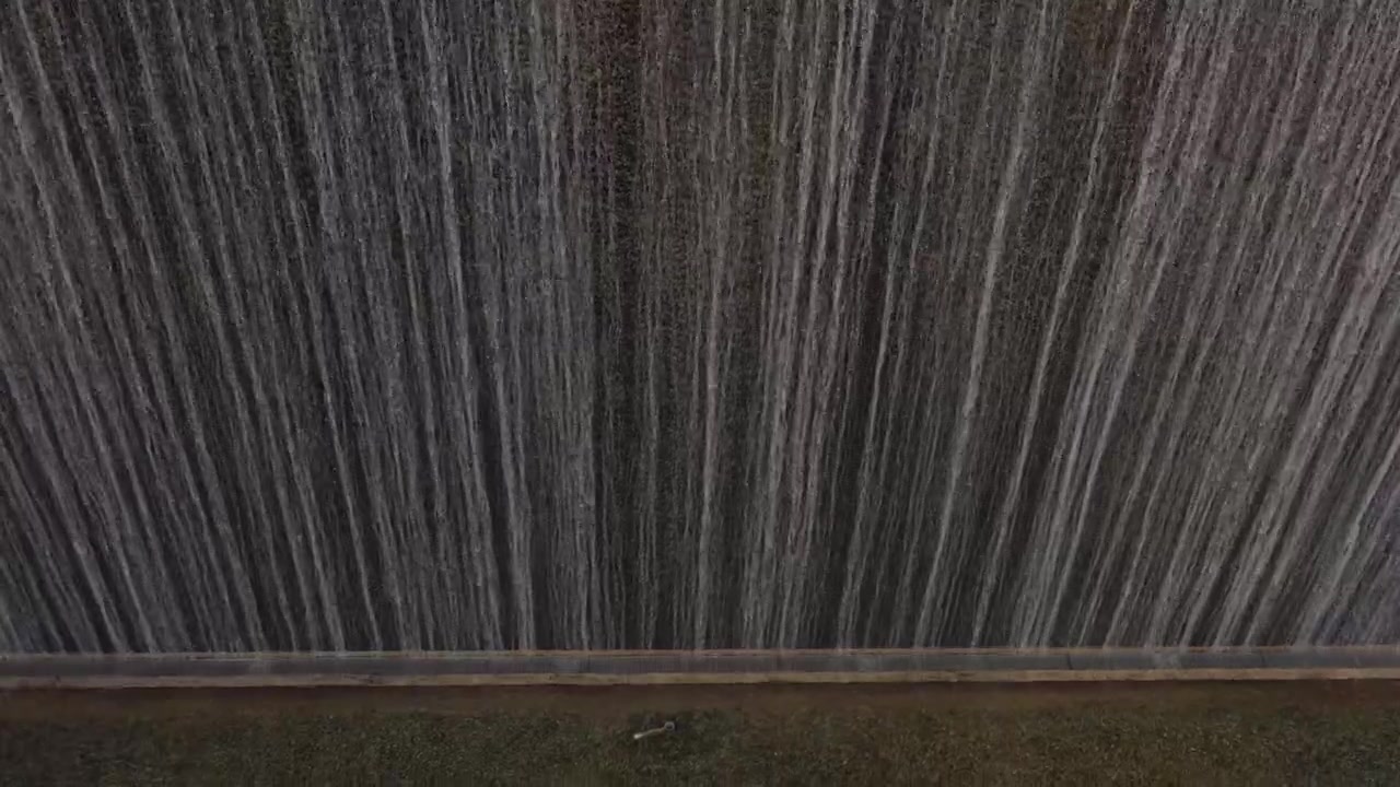 Densa cortina d'acqua che cade in striature verticali contro una superficie scura con una sporgenza alla base.