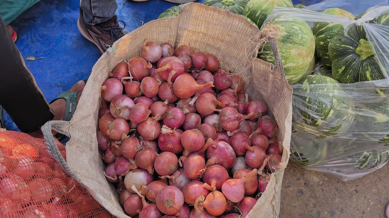 Camera circles around a sack full of red onions, showing their smooth skins and natural shine, with pumpkins and market produce nearby adding vibrant detail to the frame