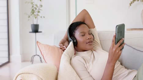 happy african american woman lying on sofa wearing headphones and using smartphone, slow motion