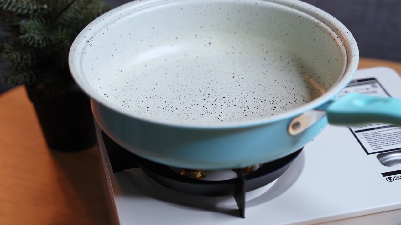 A close-up shot of a white bowl being moved near a portable gas stove with brass burners. The cooking equipment sits on a wooden table indoors beside a small potted pine plant.