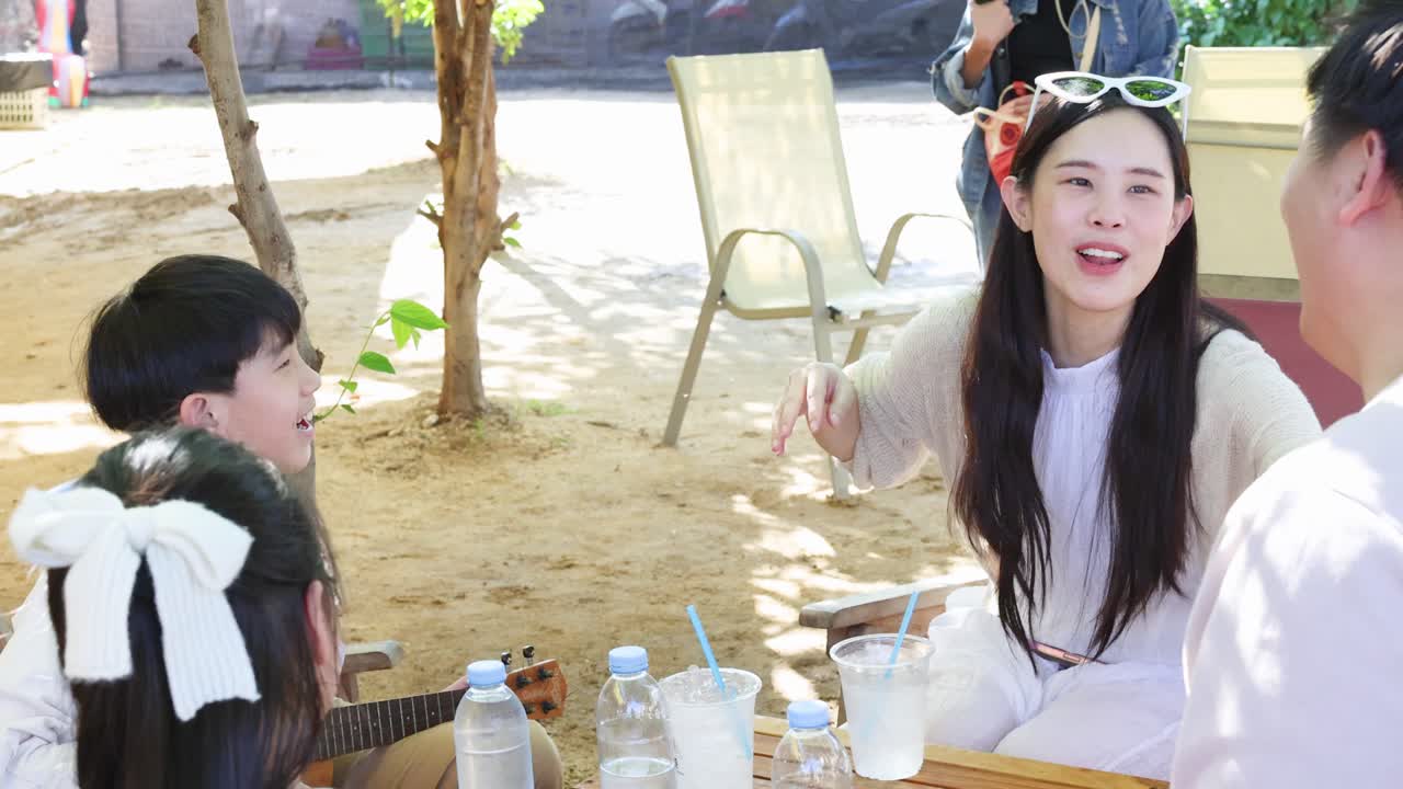 พ่อแม่และน้องสาวให้กำลังใจเด็กชายตัวเล็กกำลังฝึกเล่นอูคูเลเล่ที่โต๊ะคาเฟ่กลางแจ้งที่มีแดดส่อง