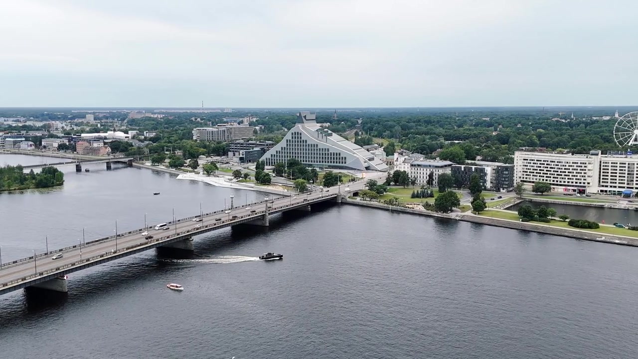 Il traffico si muove attraverso il Ponte di Pietra oltre la Biblioteca Nazionale della Lettonia mentre le barche navigano lungo l'ampio fiume Daugava a Riga.