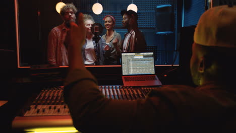 rear of the man sound engeneer sitting at the sound board in the dark studio and mastering the song while male and female four people group singing together and recording at the microphone