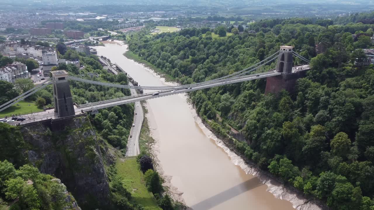 Drone video showcasing the iconic Clifton Suspension Bridge in Bristol, England, spanning the scenic Avon Gorge. The bridge architecture and lush greenery are bathed in soft daylight