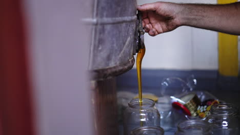 farmer-beekeeper filling glass jar with freshly extracted golden bee honey, flowing-dripping then turns off the handle of the extractor faucet with his hand, traditional honey production