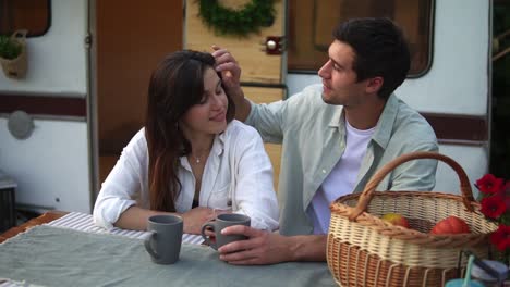 coppia allegra uomo e donna che bevono caffè seduti insieme a un tavolo di legno all'aperto. uomo che accarezza i capelli della sua amata, legame. casa con ruote stilose e statiche sullo sfondo