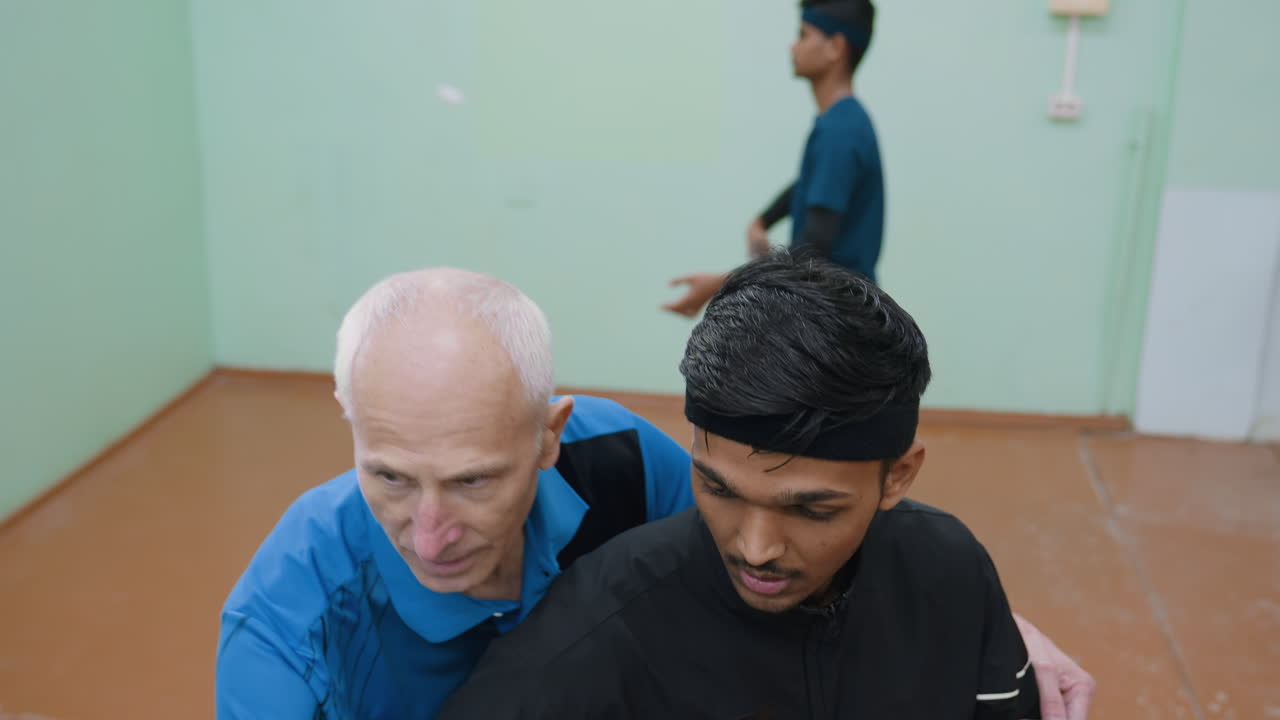 Father showing son how to maintain proper posture when serving tennis ball while another young man trains in background by hitting tennis ball against wall in indoor sports hall session