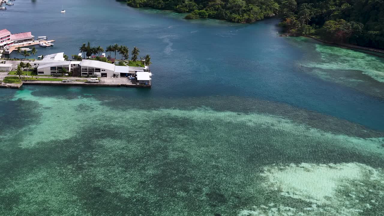Ripresa aerea con drone che cattura acqua cristallina turchese, barriere coralline e un complesso edilizio costiero.