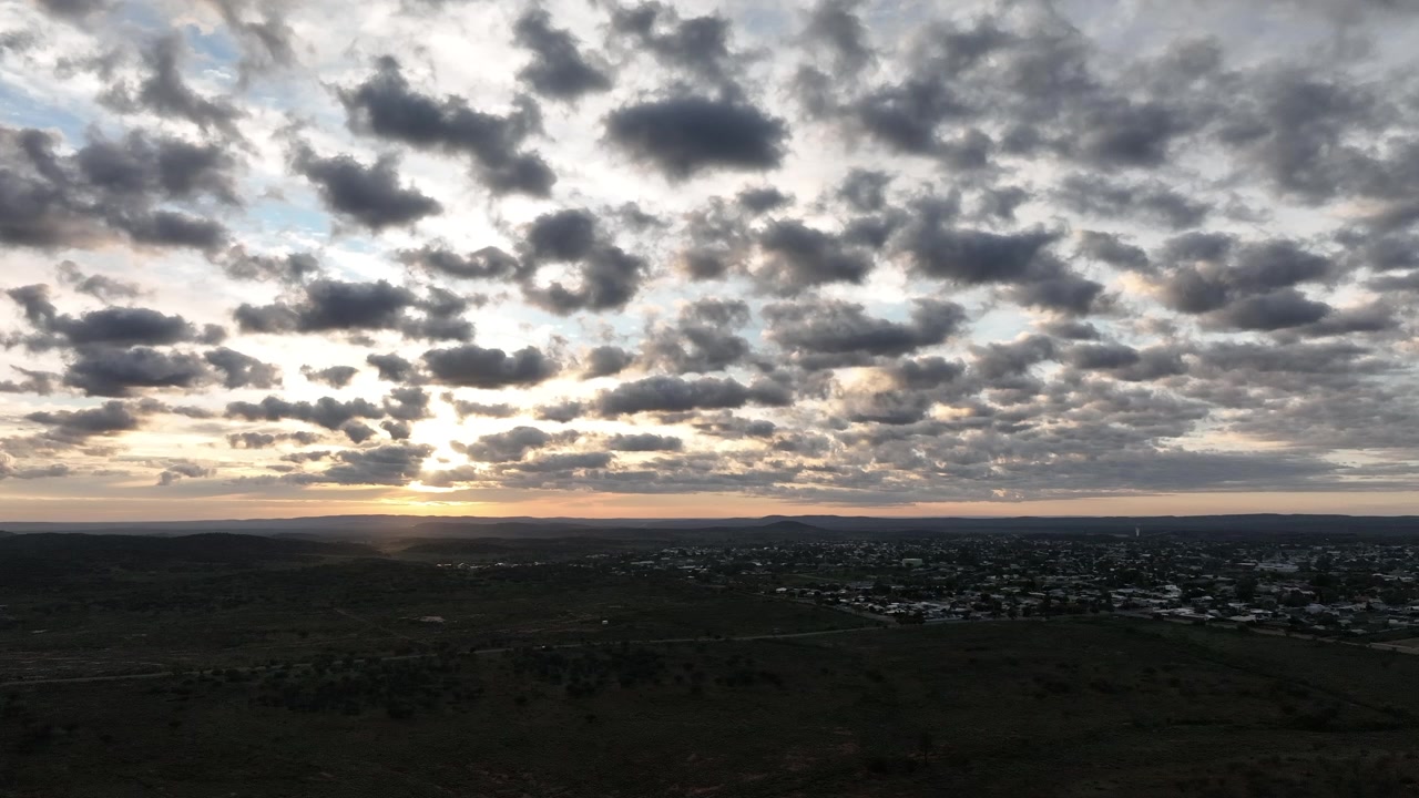 Wide aerial view of Broken Hill and the surrounding outback as the rising sun breaks through scattered clouds, casting soft rays of light across the distant landscape.