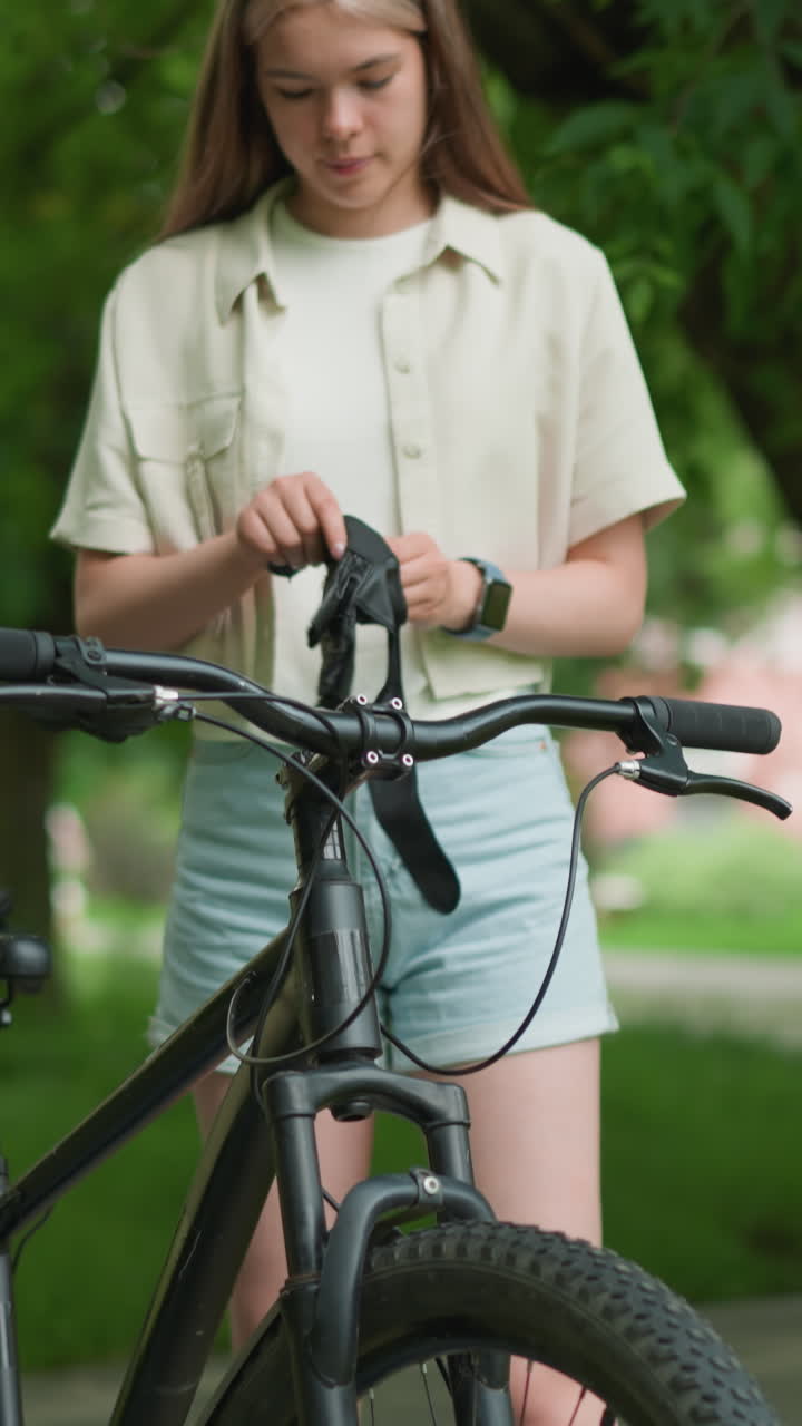 아름다운 소녀는 울창한 초록색과 나무로 둘러싸인 포장된 길에서 자전거 에 검은 장갑을 조정합니다. 건물의 흐릿한 배경과 바람에 날아다니는 잎과 함께.