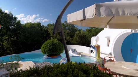 Traveling-Shot-of-a-Circular-Pool-with-a-Jacuzzi-and-a-Tree-in-the-Center,-featuring-a-White-Circular-House-and-an-Open-Umbrella-in-the-Foreground