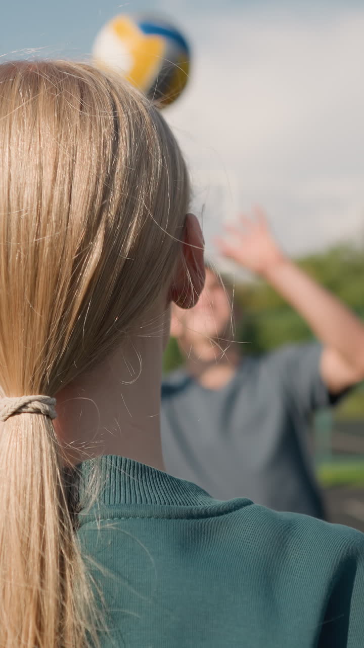 Vista posteriore di una ragazza con felpa verde e capelli legati che si allena a pallavolo con i compagni sullo sfondo, concentrata sul lavoro di squadra e sulla pratica della pallavolo in un ambiente esterno