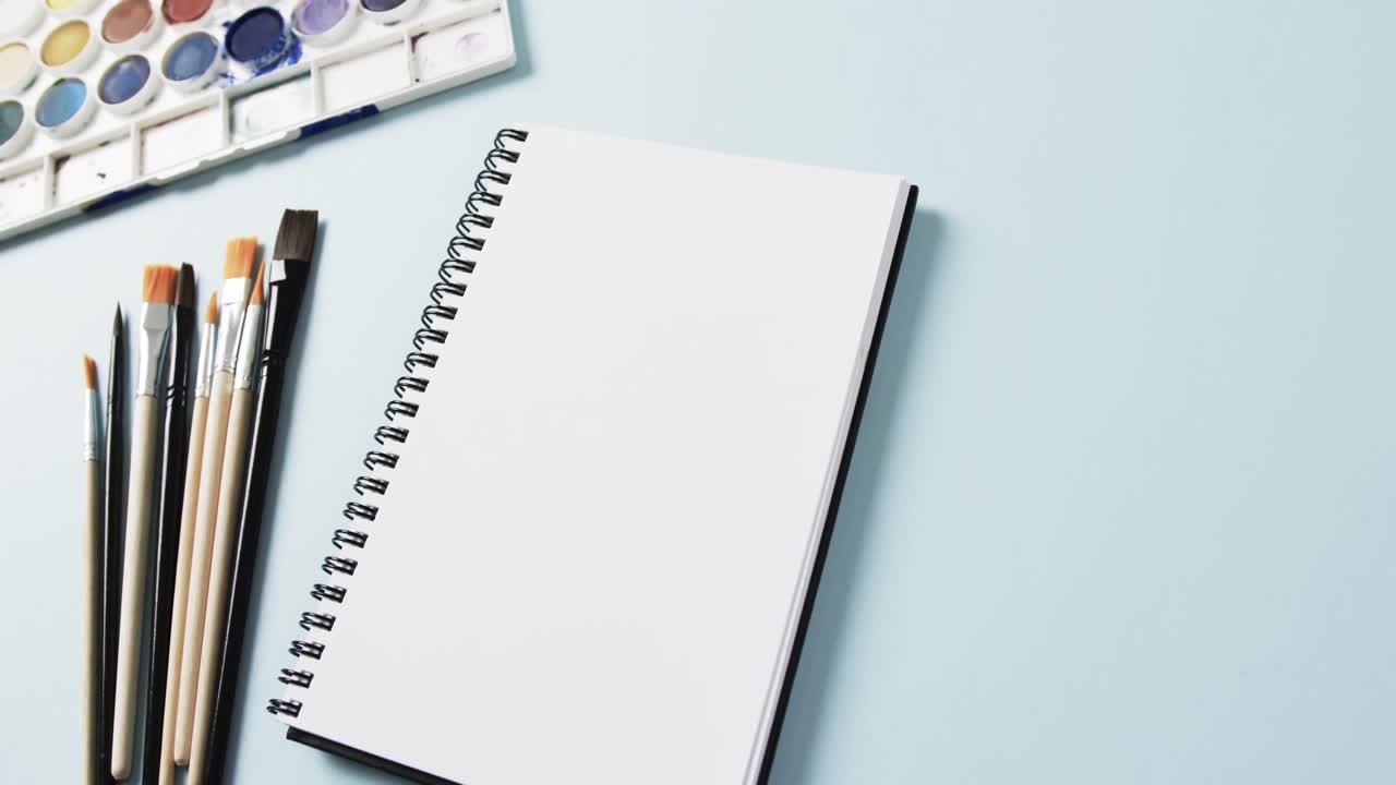primer plano del cuaderno, pinceles y pinturas sobre fondo azul, en cámara lenta