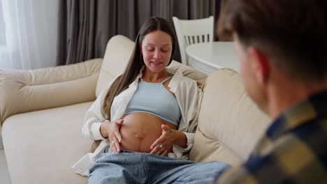 Over-the-shoulder-Happy-pregnant-brunette-woman-with-blue-eyes-lies-on-the-sofa-and-communicates-with-her-middle-aged-husband-at-home