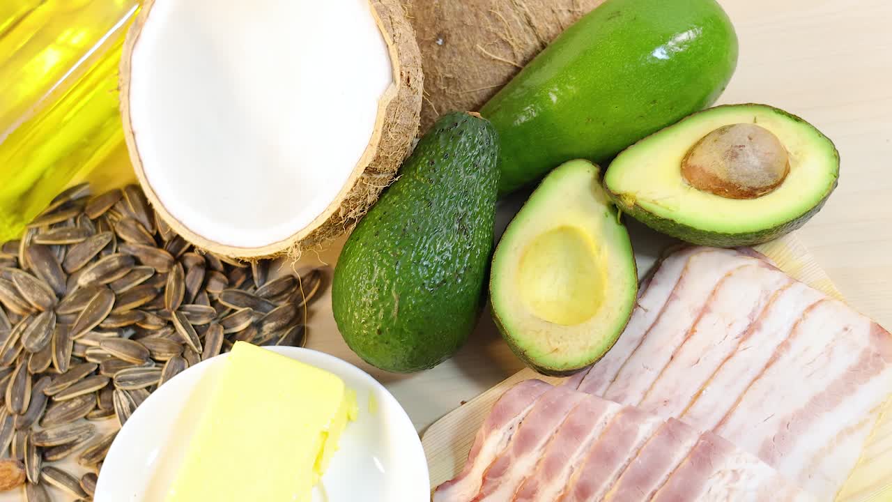 A variety of healthy fats including nuts, seeds, oils, and avocados arranged on a light wooden surface