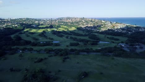 Drone-flying-over-a-rugby-soccer-field-overlooking-Bluff-golf-course-with-green-scenery-with-a-road-dividing-and-the-ocean-in-the-background