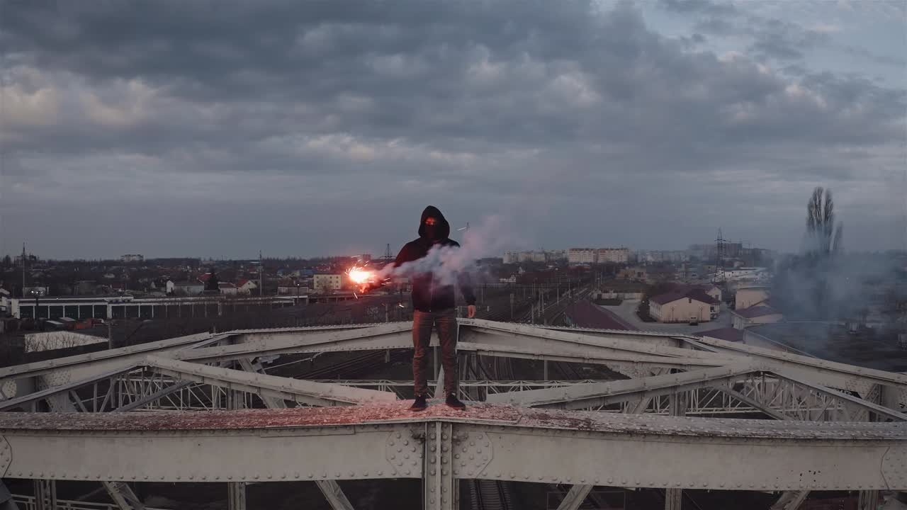 vista aérea de un joven con capucha y balaclava de pie en la parte superior de la construcción del marco de un viejo puente de marco de acero con una llamarada de señal de quema roja