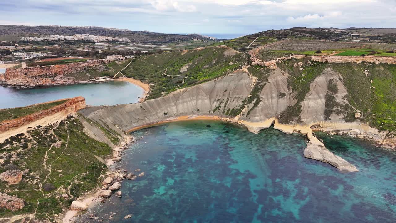 Malta'nın Mġarr bölgesindeki Qarraba Koyu, kireçtaşı platosunun altındaki mavi kil yamaçlarıyla Ġnejna Koyu sistemi içinde berrak Akdeniz suyu üzerinde kavisli bir koy oluşturuyor; aktif kıyı erozyonu ve pürüzsüz drone yörünge hareketi.