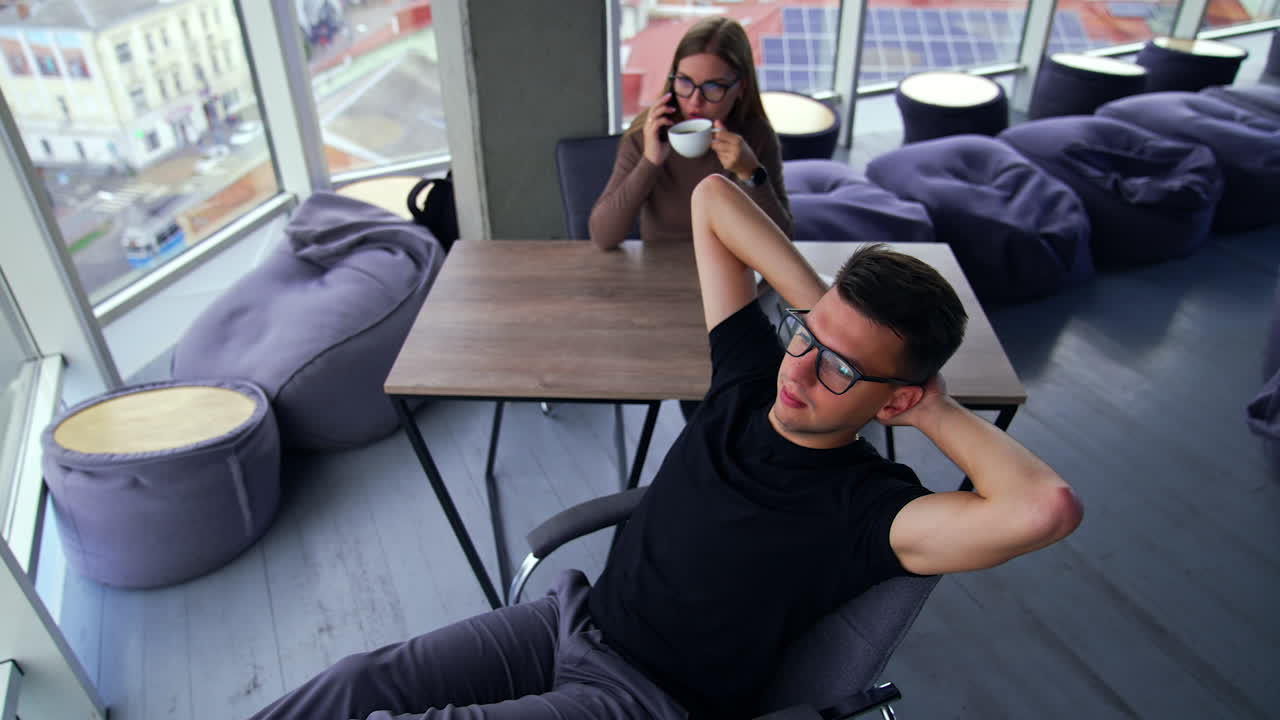Relax from work in the office. Man stretched in his chair, his legs on the table. Female colleague drinking coffee and speaking on the phone. High angle view. City view in the window.