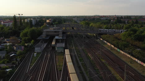 Forwards-tracking-of-train-arriving-into-station-while-tram-crossing-railway-tracks-over-Bosebrucke-bridge.-City-scene-in-evening-holden-hour.-Berlin,-Germany