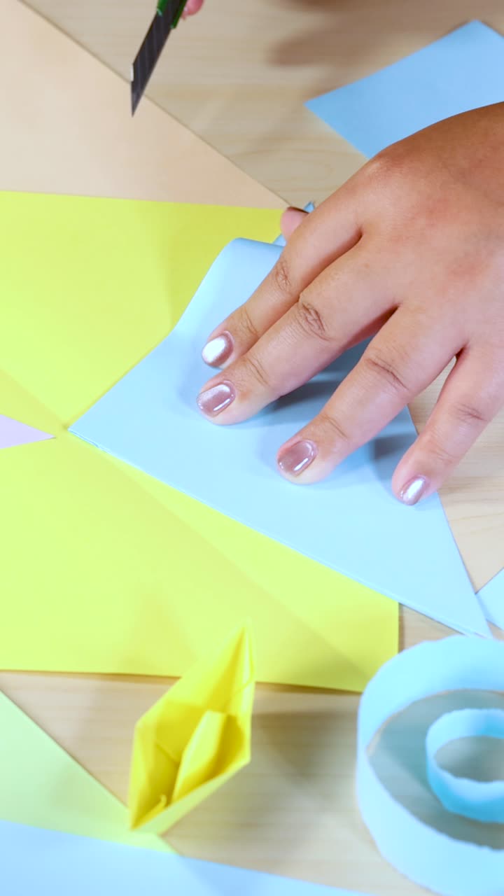 Hands use craft knife to cut colored paper on table, bright lighting, close-up perspective