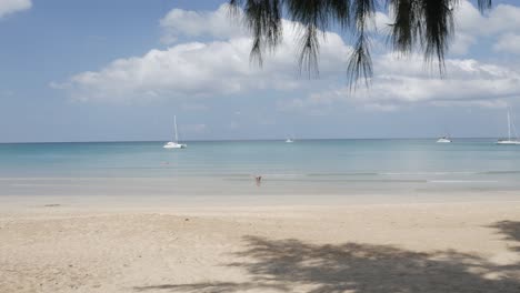 Der-Strand-Mit-Ein-Paar-Yachten-Im-Meer,-Weißer-Sand-Und-Wellen-Vom-Ruhigen-Meer-Bei-Sonnenschein-Tagsüber