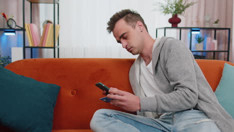 Man-sitting-on-couch-using-credit-bank-card-and-smartphone-while-transferring-money-online-shopping