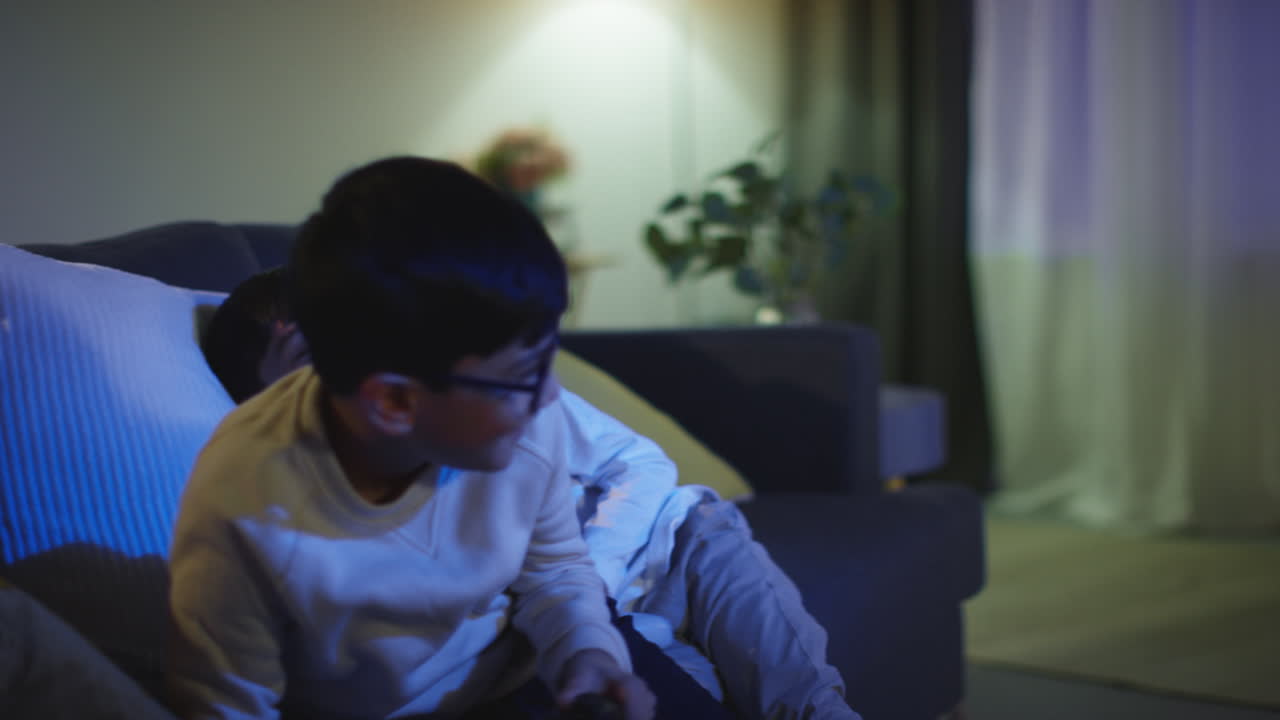집에서 두 명의 어린 소년이 컴퓨터 게임 콘솔을 가지고 재미있게 놀고 밤 7시 늦게 컨트롤러를 위해 싸우고 있습니다.