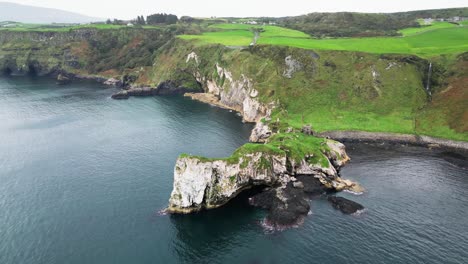 Toma-Aérea-Descendente-Frente-A-La-Costa-Del-Castillo-De-Kibane-En-Irlanda-Del-Norte-A-Lo-Largo-De-La-Carretera-Costera-De-La-Calzada-Gigante-Con-Vistas-A-Los-Acantilados-Costeros-Y-Al-Paisaje-Verde-Prístino