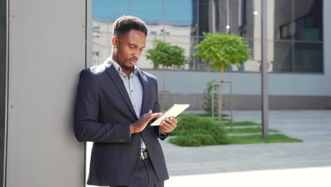 young african american businessman in formal business suit standing working with tablet in hands on background modern office building outside